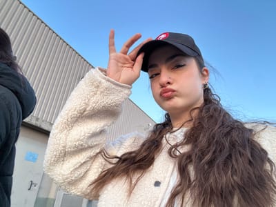 A young woman with long, curly brown hair, wearing a black baseball cap with a red and white logo and a cream-colored sherpa jacket, poses for a selfie. Her lips are puckered in a kiss. A portion of another person wearing a black coat is visible on the left. The background includes a metal building with vertical lines and a blue sky.