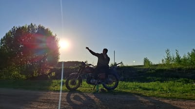 A man sits on a red motorcycle on a dirt road near Шешурга, Russia. The motorcycle is parked on a kickstand in a grassy area. A large tree is to the left of the motorcycle, obscuring the setting sun, creating a lens flare effect. The man is silhouetted against the bright sky, and is on the phone while raising his other hand. Power lines stretch across the sky in the background. Bushes and trees line the horizon.