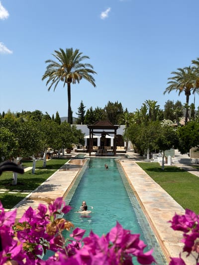 The media file depicts a serene outdoor scene with a swimming pool and lush greenery, set in Santa Eulària des Riu, Spain. The image is oriented vertically, with the sky occupying the top left portion and a long, rectangular swimming pool dominating the right side. In the pool, a person is swimming, appearing relaxed and enjoying the water.
The pool is surrounded by a light-colored tiled edge and a strip of manicured green lawn. Beyond the lawn, a pathway lined with what appear to be white statues or sculptures runs parallel to the pool. Further back, there is a dense arrangement of trees and foliage, including palm trees, contributing to a tropical or Mediterranean ambiance. A white structure with a dark roof, possibly a gazebo or covered seating area, is visible amongst the trees.
In the foreground, vibrant purple bougainvillea flowers are in sharp focus, adding a splash of color and a sense of depth to the composition. The sky is a clear, bright blue with a few wispy clouds, suggesting fair weather and a daytime setting. The overall mood is one of tranquility and leisure. No visible text is present in the image.