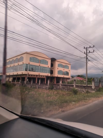 The image presents a roadside view in Bateiiliek, Indonesia, captured from within a vehicle, with reflections visible on the windshield and dashboard in the foreground. The scene is under an overcast sky, with numerous utility wires stretching across the frame, originating from a prominent wooden utility pole on the right.
The central subject is a two-story, peach-colored building featuring a distinct arched roof and multiple large, rectangular windows with white frames. The ground floor appears to be open or unfinished, with visible pillars and an empty, somewhat dilapidated aesthetic, suggesting it might be an abandoned or underdeveloped commercial property. A low concrete wall separates the building from the roadside.
In the mid-ground, beyond the main building, a house with a red roof is visible, and distant mountains or hills can be seen under the cloudy sky. The foreground shows blurred grass and sparse vegetation beside the road. No people or visible text are present in the image. The time of day is daytime, characterized by diffuse light from the cloudy conditions. The overall mood is quiet and somewhat muted due to the overcast weather and the seemingly disused building.
