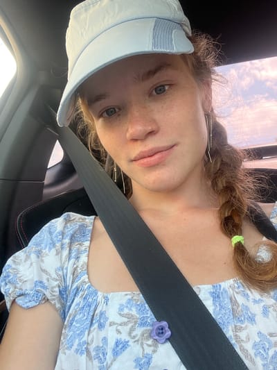A young woman with reddish-brown hair braided and a light blue cap sits in the passenger seat of a car, wearing a blue and white floral print top and a seatbelt. She has freckles on her face and is wearing long, dangling earrings. A light purple flower-shaped button is visible on her dress, partially obscured by the seatbelt. The background shows a blurred view of the sky with clouds, suggesting the car is in motion. The interior of the car, including the black seat and seatbelt, is also visible. The scene appears to be outdoors, possibly on a road.