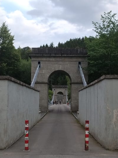 The image shows a pedestrian walkway leading towards a suspension bridge. The walkway is flanked by light-colored concrete walls. In the foreground, two red and white striped bollards are visible on the right side of the walkway. The walkway leads to a large stone archway that is part of the bridge's support structure. Metal cables are visible descending from openings in the stone structure. In the background, more of the suspension bridge and the surrounding landscape of trees are visible. A few people are walking on the bridge in the distance. The sky is cloudy. This is the Rope Bridge in Slavonice, Czechia.