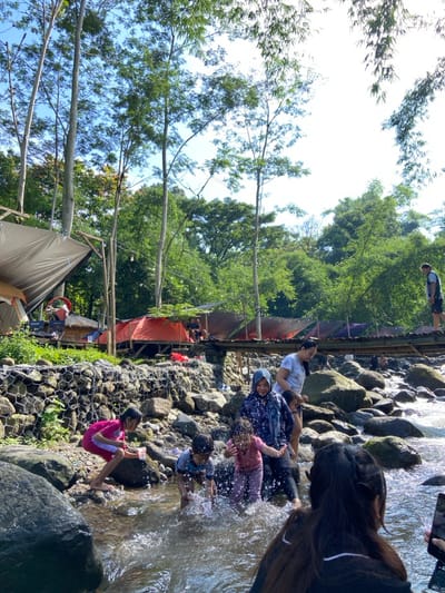 A daytime shot shows a group of people enjoying a rocky creek. Adults and children sit and splash in the water. A tent and a small building with a red tarp are visible on the left bank, along with trees and vegetation. A person with long hair stands near the water's edge, possibly taking photos with a phone. A person is walking on a structure in the distance.