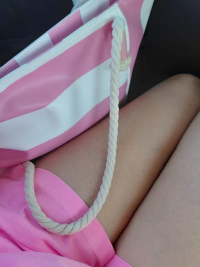 The image shows a close-up view of a person's legs and a pink and white striped bag. The bag appears to be made of a canvas-like material and has a thick, braided rope handle. The person is wearing bright pink shorts. The background is dark and out of focus, suggesting it might be an indoor setting or a car interior. There is no visible indication of the location being in Aqsu city administration, Kazakhstan.