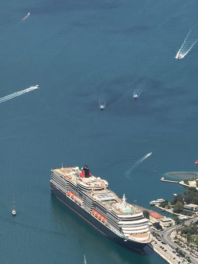 An aerial image provides a high-angle view of a large cruise ship near a coastal town, likely in Špiljari, Montenegro. The massive ship, featuring multiple decks, numerous lifeboats along its side, and a distinctive red and black funnel, occupies a significant portion of the lower right frame, appearing either docked or moored very close to land.
The calm, deep blue water around the cruise ship is bustling with activity from several smaller boats. These are mostly speedboats, each leaving a clear white wake, indicating movement. At least five such boats are visible; some are white, and one distinctively red/orange boat is seen in the upper left. A red and a yellow/orange buoy are also present in the water.
The coastline, visible in the upper right, features a developed area with a winding road, numerous parked cars, and various buildings, many with red-tiled roofs, nestled among green trees. A small, enclosed harbor or marina with additional small vessels is also part of the coastal landscape. The scene is bathed in bright daylight, suggesting clear weather. No visible text is discernible on any of the objects or structures within the image.