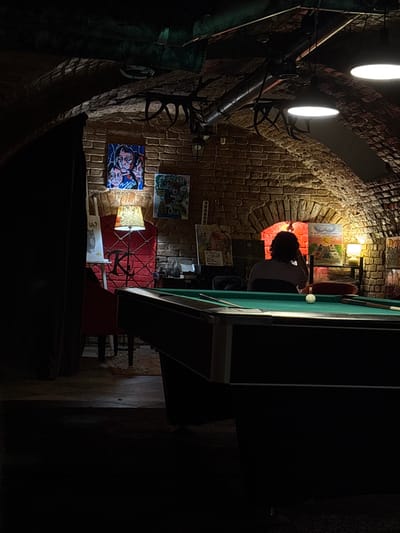 The image shows a dimly lit, arched brick room, possibly a bar or recreational space, in Svkh Maslovskiy 1 Otdeleniye, Russia. A pool table with a white ball and two cues is in the foreground. In the background, a person is seated facing away from the camera, looking towards a brightly lit arched opening with red light emanating from it. Several pieces of artwork are visible on the brick walls, including a triptych-like painting above a red chair with a patterned lampshade. The ceiling features exposed pipes and industrial-style lighting fixtures. The overall atmosphere is moody and artistic, with a focus on the interplay of light and shadow.