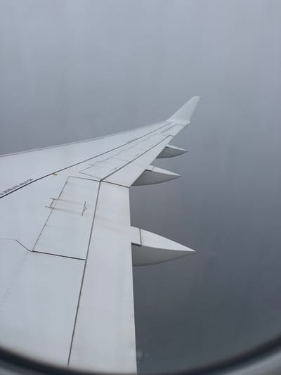 A forensic description of the scene depicts an aerial view from an aircraft passenger window, showcasing the right wing of a commercial airliner in flight, identified as being over Baambrugge, Netherlands. The white wing occupies the left and central portions of the frame, extending towards the upper right, with visible panel lines, retracted flap segments, and three engine fairings (nacelles) positioned beneath. Dark, linear smudges, consistent with exhaust residue or dirt, are present on the wing's upper surface, specifically over an inboard flap section. Partial text, reading "NO STEP OUTSIDE T", is visible on the wing's leading edge area. The environment beyond the wing consists of an unbroken, uniformly grey and overcast sky above, transitioning to a dark, indistinct land or water mass below, obscured by atmospheric haze and distance. The curved edge of the aircraft window frame is visible at the bottom of the image. No individuals or specific ground landmarks are discernible within the visible area.