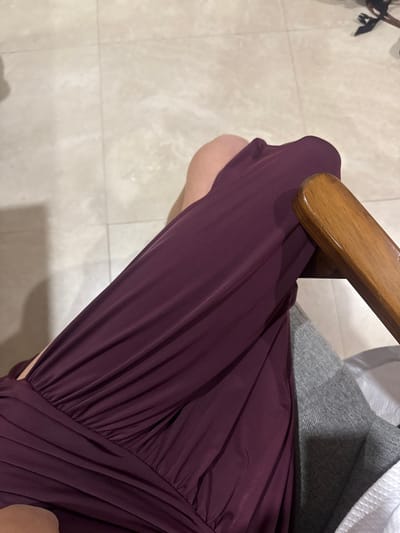 A top-down shot reveals a seated person wearing a flowing, wine-colored dress. The fabric drapes and folds elegantly, creating a sense of movement. The person is seated on a chair with a light wood armrest and a grey cushion. A tiled floor provides a cool-toned contrast to the rich color of the dress. The composition focuses on the texture and color of the dress, drawing the eye along its length.
