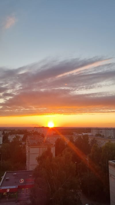 The image captures a sunset over the city of Aleksin, Russia. The sky is a gradient of blue at the top, transitioning to hues of orange and yellow as it approaches the horizon. Wispy clouds are present, reflecting the sunset's colors. A bright sun is visible, partially obscured by clouds, casting a warm glow across the sky and illuminating the urban landscape below.
The foreground features a cluster of apartment buildings with flat roofs, characteristic of Soviet-era architecture. The buildings are predominantly light-colored, with visible windows and balconies. Some trees, with dense green foliage, are interspersed among the buildings, some reaching up towards the lower branches of the buildings. A commercial building with a red and white facade and a prominent circular red logo with a white "5" is situated in the lower left portion of the image. Lens flares from the setting sun streak across the scene, adding a dramatic effect. The overall impression is of a quiet, residential area at the end of the day.