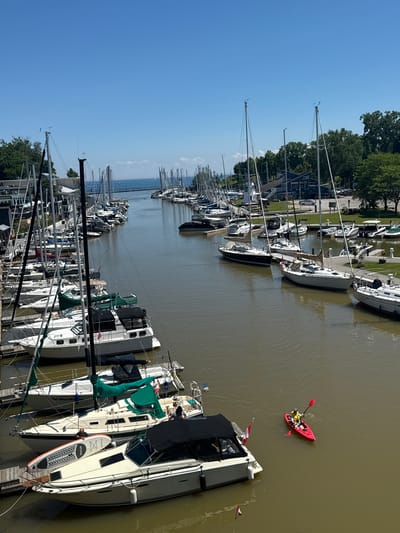 This is a high-angle, full-shot, eye-level, outdoor, daytime, vertical, color photograph of a marina in Oakville, Canada. The marina is filled with numerous sailboats and motorboats docked side-by-side, creating a dense urban maritime scene. The water is a murky brown, reflecting the overcast sky. In the foreground, a red kayak with a single occupant wearing a yellow life vest and a white helmet navigates the waterway, holding a red paddle. Several large sailboats are docked along the left side of the frame, with their masts reaching high into the clear blue sky. To the right, more boats are visible, as well as a building with a blue facade and white trim, likely a marina office or restaurant. Trees line the background, with a few more boats visible in the distance, suggesting a larger harbor. The overall impression is one of a busy recreational boating area on a clear day.