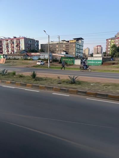 The image displays an urban street scene in Biashara ward, Kenya, under a clear blue sky. In the foreground, a multi-lane asphalt road features a central median bordered by yellow and black curbs, containing sparse vegetation and several agave-like plants.

On the far side of the road, a white compact car with a damaged rear window is parked facing right. A male individual, dressed in dark clothing, walks left to right on the road's shoulder, passing a tall street light pole. Further right, a person on a motorcycle also travels left to right.

The background is dominated by multi-story buildings. To the left, a large residential building with red balconies and gray walls is prominent. Behind it, a smaller structure bears a sign for "Redeemed Gospel Church Inc." A central multi-story building with gray walls and green accents features water tanks on its roof. Another multi-story building, partially obscured, is visible to the right, also with rooftop water tanks, alongside an apartment-style building with green and gray sections on the far right.

Two billboards are present: a green one displaying "Safaricom m_pesa" and a white one partially reading "NOT FOR SALE" and "ELVAC AUTO REPAIR." Utility poles and overhead power lines are visible. Several indistinct human figures are present near the buildings in the mid-ground. The scene depicts an active daytime urban environment.