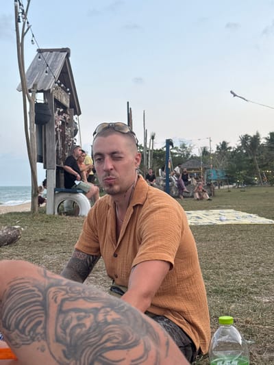 The scene depicts an outdoor gathering on a grassy area adjacent to a beach, identified as being in Ban Pang Pring, Thailand, during daylight hours under a light blue sky with sparse clouds.
In the foreground, a male with a shaved head and sunglasses perched on top, wearing an orange collared shirt, winks his right eye and pouts his lips, looking directly towards the viewer. Tattoos are visible on his left forearm. An out-of-focus, heavily tattooed arm belonging to another person is in the extreme lower-left foreground, partially obscuring the main subject. To the main subject's right, a clear plastic water bottle with a green cap rests on the grass.
In the midground-left, a sandy beach meets the ocean, with small waves visible. A rustic wooden structure, possibly a hut or decorative shrine, featuring a pointed roof and string lights, stands near the water's edge. A male with dark hair and a beard is seated on a white tire that serves as a bench base next to this structure.
In the midground-right and background, a large grassy expanse is occupied by multiple individuals seated or standing casually. A patterned yellow and white mat or blanket is spread on the grass. Various wooden poles and structures are scattered throughout this area, some appearing as decorative elements or informal seating. Further back, a small, light-colored hut with a dark roof is visible among tropical trees, including palm trees, which line the distant horizon. High in the sky on the far right, a light-colored kite with a long tail is visible.