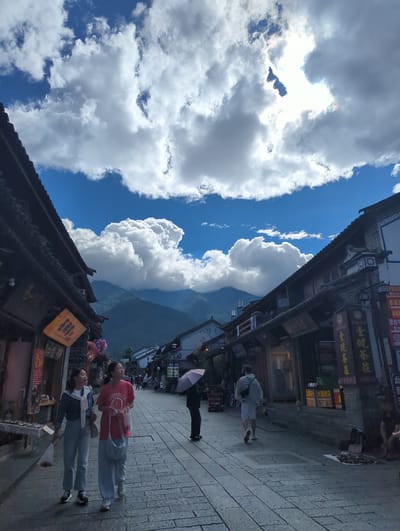 The image depicts a street scene in Dali City, China, under a partly cloudy sky. The street is paved with stone tiles and lined with traditional two-story buildings with dark tiled roofs. Several people are walking along the street. In the foreground, two young women are walking towards the viewer. The woman on the left is wearing a blue jacket over a white shirt and light blue pants. The woman on the right is wearing a red t-shirt and light blue pants. Both are carrying bags. Further down the street, a person holding a pink umbrella is walking away from the viewer. In the background, mountains are visible. Shops line both sides of the street, with signs in Chinese characters. The overall atmosphere is one of a bustling but relaxed tourist area.