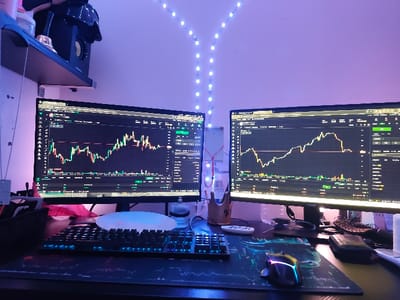 The image shows a desk in a room located at 32.314 latitude, 34.941 longitude in Kfar Yona, Israel. Two computer monitors display a stock market chart with fluctuating values. There are two computer mice on the desk, a keyboard, a cell phone, and a black speaker. The room is lit by a string of white LED lights. The wall is painted a light purple color. There are no people visible in the image. The scene appears to be a home office or a personal workstation. The image captures a moment in time, showcasing the current state of the stock market chart.