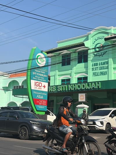 The image captures a street scene in Pekalongan, Indonesia, dominated by the green facade of RS Siti Khodijah. A large, curved green and white sign stands prominently in the foreground, displaying the hospital's name in both Indonesian and Arabic script. Text on the sign indicates "Rumah Sakit Siti Khodijah," "IGD 24 JAM," and the tagline "Melayani karena Allah." Below this, contact information including phone numbers and an address in Pekalongan are visible.

A man is riding a black motorcycle on the street, wearing a black jacket, an orange t-shirt, and denim shorts. He is also wearing a helmet. In the background, several cars are parked or driving on the road. A dark gray SUV is visible on the left, and a white car with license plate "1054 RA" is on the right. The building itself features arched windows and architectural details consistent with modern Indonesian public buildings. The sky is clear and blue, and power lines crisscross the upper portion of the image.