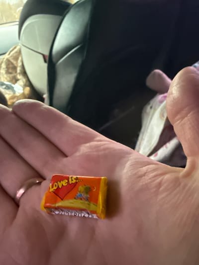 A hand is holding a wrapped piece of "Love is..." chewing gum. The wrapper is orange and features a cartoon illustration. A ring is visible on one of the fingers. In the background is a dark object. There is no information that refers to the city of Klenovskoe, Russia.