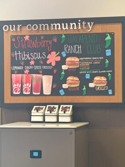 Here is a forensic description of the image:
The image presents a restaurant menu board displaying food and beverage offerings. The board advertises a "Strawberry Hibiscus" drink made with lemonade, Sunjoy, Sprite, and frosted variations. It also showcases a "Jalapeno Ranch Club" sandwich, available in spicy, regular, and grilled versions, which features buttermilk ranch bun, caramelized bacon, pepperjack cheese, chicken, tomato, lettuce, and pickled jalapenos, served with creamy jalapeno ranch.
The menu is mounted on a wooden and blue frame with the words "our community" affixed to the frame. Next to the menu board is a condiment station with small containers labeled "Mint" and "TO" (likely a shortening of tomato). A grey food warmer or holding cabinet with a "Ready" indicator light is positioned next to the condiment station.
Given the menu items and drink designs, it is highly probable that this image was taken inside a Chick-fil-A restaurant located somewhere in the United States of America. The absence of any landmarks or street signs in the frame prevents a definitive geographic location within Mountain Park, United States of America, to be determined.