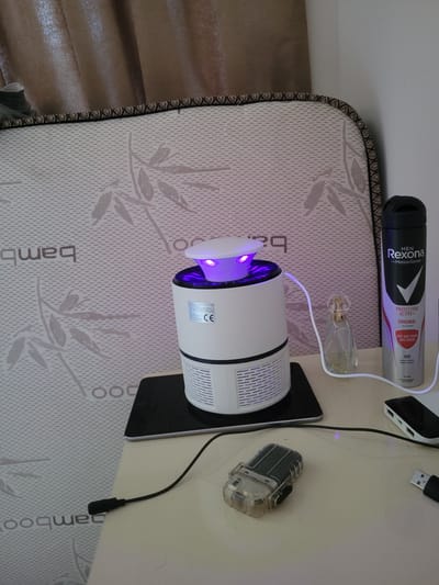 The image shows a close-up view of a bedside table with various objects. In the background, a mattress with a bamboo pattern is visible, along with brown curtains. The objects on the table include a white mosquito repellent device with purple LED lights, a small glass perfume bottle, and a can of Rexona Men deodorant. There are also charging cables and a small transparent case containing what appears to be a lighter. The scene is indoors, and there is no indication of the city of Milisi, Georgia.