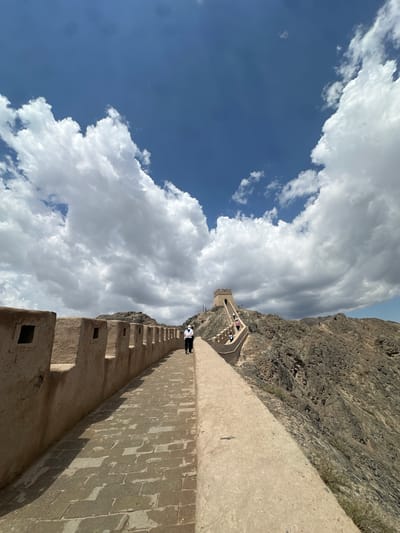 A wide-angle, eye-level, outdoor photograph captures a section of the Great Wall of China, likely near Jiayuguan, China, under a bright, partly cloudy blue sky. The wall, constructed from tan-colored bricks and stone, snakes along a rugged, rocky mountainside. The walkway on the wall is paved with rectangular stones. Along the top of the wall are crenellations, with square openings visible.
In the foreground, a person wearing a white shirt, dark pants, and a white face mask walks away from the camera along the wall. Further in the distance, the wall ascends a steep incline towards a fortified watchtower situated atop a rocky peak. Several other individuals can be seen on the wall pathway and climbing towards the watchtower. The surrounding landscape is characterized by barren, rocky hills and sparse vegetation. The lighting suggests it is daytime, with the sun casting shadows on the pathway.