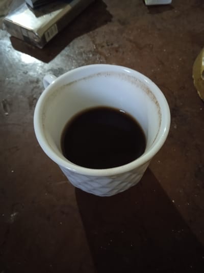 A close-up, high-angle shot shows a white mug filled with dark liquid, presumably coffee, placed on a dark, textured surface. The mug has a quilted pattern on its exterior and a faint ring of residue is visible along the inside rim. In the upper left corner of the frame, a portion of a cigarette pack is visible. The background is out of focus, but appears to be a table with some scattered light-colored specks. The scene is lit, creating shadows. There are no people present. The image does not provide any specific information about the location of Aznvadzor, Armenia.