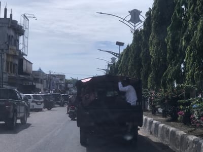 This daytime image captures a street scene in Kutalhoksukon, Indonesia, seemingly taken from a moving vehicle. The sky is overcast, contributing to a soft, diffuse light over the area.

In the foreground, a dark-colored pickup truck or similar utility vehicle is central, viewed from behind. Several people are riding in its open bed; one individual in a white shirt is standing on the right, holding onto the roof frame. To the left of this truck, a motorcycle with a rider in a helmet and light head covering is visible.

The road ahead shows other vehicles, including a black pickup truck on the far left, followed by a white minivan (with license plate "BL 3559 RC") and several dark cars. A motorcycle's license plate, "BL 6756 KBA", is also visible. Traffic appears to be moving or queuing.

On the left side of the road, multi-story buildings with traditional architecture, featuring arched windows and balconies, line the street. A large, empty metal frame, possibly for a billboard, extends from the side of one building. Faint text on signs above storefronts includes "SINAR FOTO" and "POS KUE".

The right side of the road is characterized by a long row of tall, dense green trees, creating a lush wall of foliage. Interspersed among these trees are decorative streetlights with ornate, crest-like fixtures and solar panels on top, suggesting modern but aesthetically designed infrastructure. A concrete curb separates the road from the planted area, where some pink flowering bushes can also be seen. The scene depicts a busy urban or semi-urban environment.