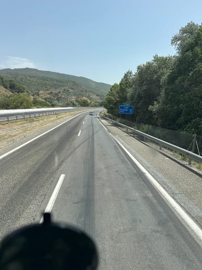 The image captures a forward view from a vehicle on a highway in the vicinity of Cambil, Spain. The road, an asphalt surface, is marked with white lines and tire tracks are visible. To the right, a metal guardrail separates the road from a gravel shoulder and a chain-link fence. Beyond the fence, dense green trees line the road. A blue road sign indicates "E-902" and "A-44" with destinations "Granada 58" and "Motril 133", suggesting the location is on the A-44 highway. In the background, rolling hills covered in vegetation and some scattered olive trees are visible under a clear blue sky. A white car is seen driving ahead on the highway. A dark, shadowed object, likely part of the vehicle from which the photo is taken, is visible in the lower foreground.