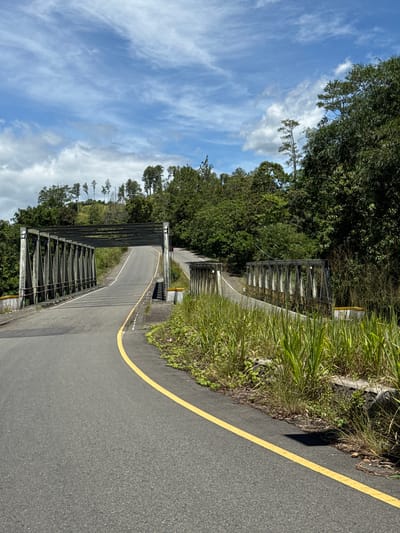 The image depicts a paved road curving through a lush, green landscape, likely in the city of Sukamulia, Indonesia. A metal truss bridge spans across the road, with another section of a similar bridge visible further along the curve. The road is marked with a bright yellow line. Dense vegetation, including tall grasses and various trees, lines the sides of the road and covers the surrounding hills. The sky above is a bright blue, with scattered white clouds and wispy cirrus clouds. The scene appears to be a rural or semi-rural setting, given the presence of the bridges and the natural, overgrown environment. There are no people visible in the image. The overall impression is one of a scenic, somewhat remote location.