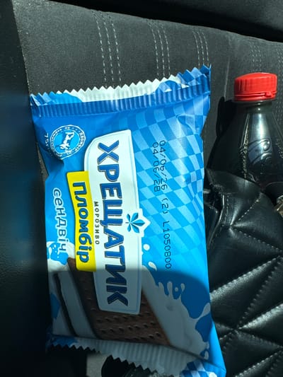 The image shows an ice cream sandwich package with Cyrillic text. The package is blue and white with a checkered pattern and graphics of milk splashes. The text on the package includes "ХРЕЩАТИК", "ПЛОМБІР", and "СЕНДВІЧ". There are also dates and codes printed on the package: "04/06/26 (21) L1050800". A red-capped bottle is visible to the right of the ice cream package. The background appears to be the interior of a vehicle, possibly a car seat made of dark textured material. The lighting suggests an outdoor environment, with sunlight casting shadows. The ice cream sandwich is the primary subject, positioned vertically in the frame. The location is identified as Sarny, Ukraine.