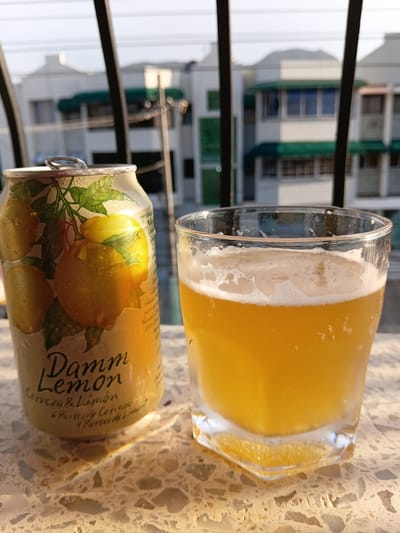 The image shows a can of "Damm Lemon" beer and a glass of beer on a terrazzo-patterned surface, likely a table. In the background, there are buildings with green awnings and white walls, suggesting a residential or commercial area. The lighting indicates it is daytime, possibly late afternoon due to the warm tones. The presence of condensation on the can and glass implies the beverages are cold. The overall scene is relaxed and casual, consistent with an outdoor setting. The image quality is good, with the subjects in focus and the background slightly blurred. The setting is likely a balcony or patio with a view of nearby structures. This specific location in Puerto Plata, Dominican Republic, could be a home, a restaurant, or a bar with an outdoor seating area.