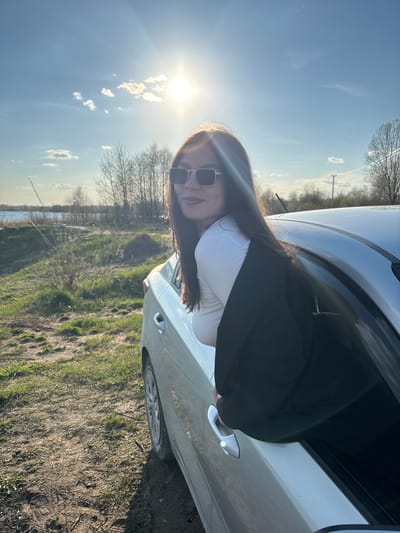 A woman with long, dark hair and sunglasses is sitting inside a light-colored car with her upper body leaning out the window. She is wearing a white top and a black blazer. The car is parked on a grassy field next to a body of water. The sun is shining brightly in the sky. The location is Zaosinovo, Russia.