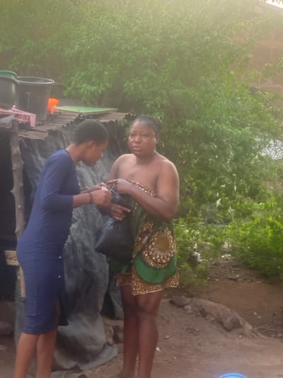 The image depicts two women outdoors in Idiroko, Nigeria. On the left, a woman wearing a dark blue long-sleeved dress and a beaded bracelet on her left wrist is observed reaching into a black plastic bag. This bag is held by a second woman on the right, who is topless and clad in a green, brown, and yellow patterned fabric wrapper covering her lower body. The second woman has braided hair and maintains direct eye contact with the viewer. Behind the woman on the left, a makeshift lean-to structure made of corrugated metal sheets is visible, supporting several plastic buckets and containers. The ground beneath the figures is bare earth, and the background consists of dense green foliage, with hints of a distant building structure through the trees. The scene is illuminated by natural light.
