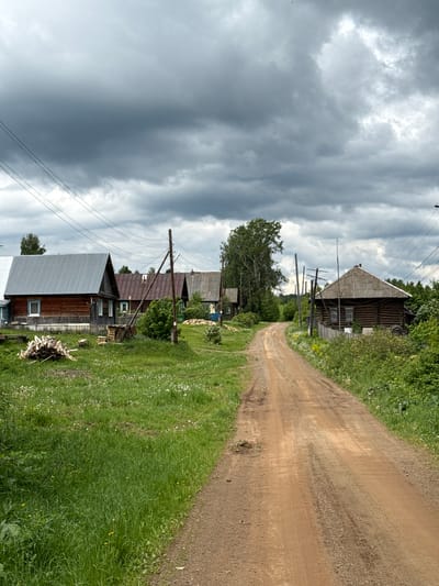 The image depicts a rural scene in Medvedka, Russia. A dirt road winds through a village, flanked by green grass and vegetation. Wooden houses line the road, constructed with traditional Russian designs, some with metal roofs. Overhead power lines run alongside the road. The sky is filled with dark, stormy clouds, suggesting an overcast day. No people or animals are visible in the scene.