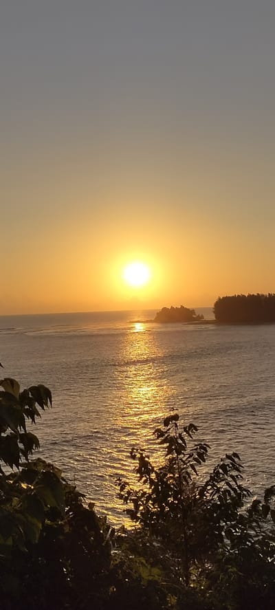The image depicts a tranquil sunset over the ocean, likely near Crakmong, Indonesia. The sun, a bright, luminous orb, is positioned near the horizon, casting a warm, golden glow across the sky and the water. The ocean surface is gently rippled, reflecting the sun's light in a shimmering path. In the distance, a small, tree-covered island or peninsula juts out into the sea, silhouetted against the vibrant sky. The foreground is partially obscured by the dark, leafy branches of trees, framing the scene and adding depth. The overall atmosphere is peaceful and serene, characteristic of a tropical coastal sunset.