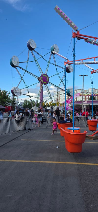This is a photograph of a carnival ride, possibly located in Pickering, Canada. In the foreground, an asphalt ground surface is marked with yellow lines. A white metal fence runs horizontally across the middle ground, separating the viewer from a crowd of people. To the right of the fence, several bright orange carnival ride buckets are visible, with a few children sitting in them. In the background, a Ferris wheel with purple and green accents is prominently displayed, with a circular sign in the center reading "Rock-O-Plane". Above the Ferris wheel, a blue sky with scattered clouds can be seen. Further in the background, buildings and trees are visible. The overall scene depicts a lively outdoor event, likely a fair or amusement park.