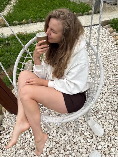 A young woman with long, wavy brown hair is sitting in a white woven swing chair outdoors. She is wearing a white hooded sweatshirt and dark brown shorts. The woman is holding a paper coffee cup to her nose and appears to be smelling it. Her eyes are closed. The swing chair is suspended by thick ropes. The ground around the chair is covered in small white and gray stones. In the background, there is green grass and some larger rocks. A wooden post is visible to the left of the swing chair. The scene appears to be in a garden or outdoor recreational area. The location is identified as Ryzhkovo, Russia.