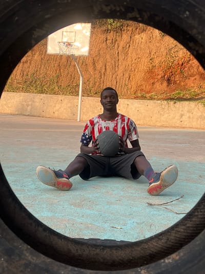 A young man wearing a red, white, and blue patterned t-shirt and black shorts sits cross-legged on a light blue basketball court, holding a dark gray basketball. He is positioned in the center of the frame, looking directly at the camera with a slight smile. His legs are extended in front of him, and he is wearing dark sneakers with red accents. Behind him, a basketball hoop with a white backboard and orange rim is visible against a backdrop of a dirt embankment covered in sparse green vegetation. The court appears to be outdoors, possibly in a rural or semi-rural area, with a concrete retaining wall running along the edge of the embankment. The foreground is partially obscured by a large, dark, circular object, possibly a tire, which frames the scene. The lighting suggests it is daytime, with the sun casting warm tones on the embankment. The location of the basketball court is in Kamakwa/Mukaro Ward, Kenya.