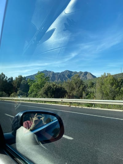 A view from inside a vehicle, possibly a car, shows a highway landscape. The asphalt road is visible on the right side of the frame. A guardrail separates the road from a mountainous terrain covered with green trees and shrubs. In the distance, a tall mountain is visible against a blue sky streaked with wispy clouds.
The car's side mirror reflects the interior of the vehicle, where a person wearing a red shirt can be seen, seemingly holding a cell phone or similar device in their hands.
Based on the mountain landscape and vegetation, this image was probably taken on the C-55 road in Esparreguera, Spain. The mountain in the background is likely part of the Montserrat mountain range.