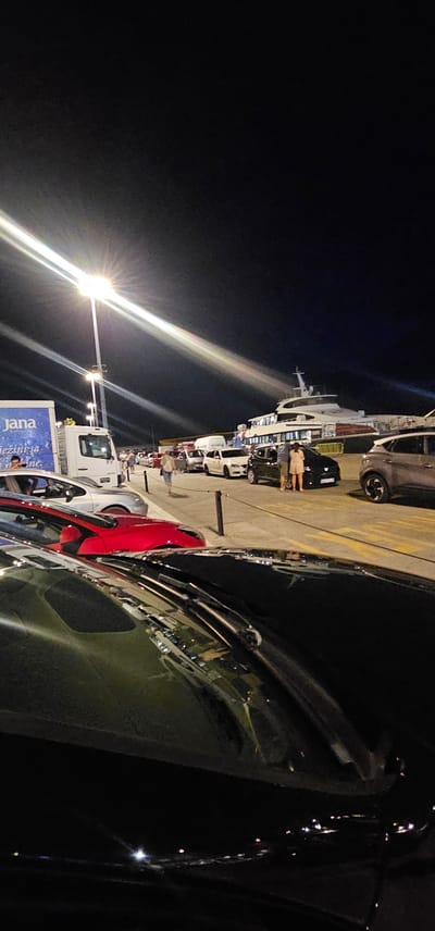 The image depicts a nighttime scene at a ferry port in Split, Croatia. In the foreground, the hood of a black car is visible, reflecting the surrounding lights. Behind it, a red car is parked, followed by a line of other vehicles including white and black cars, and a white delivery truck. The truck has the logo "Jana" on its side.
Further back, a large white ferry is docked. Several people are walking and standing near the vehicles and the ferry. The scene is illuminated by bright streetlights that cast long beams across the pavement. The sky is dark, indicating it is nighttime. The overall impression is of a busy port with vehicles waiting, possibly to board the ferry.