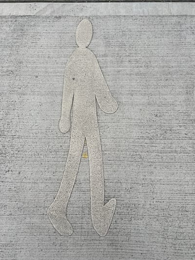 A schematic representation of a walking person is imprinted on a textured, grey concrete surface. The figure is outlined in a lighter shade of grey, with a stippled texture within the outline. The figure faces towards the right of the frame, with its left leg extended forward and its right leg back. The left arm is bent at the elbow and extends forward, while the right arm is bent and angled towards the body. The surface it is imprinted on appears to be a sidewalk or pedestrian pathway. The image is captured from a top-down perspective, showing the texture of the concrete with its horizontal striations. There is no indication of the specific location within Balbriggan, Ireland.