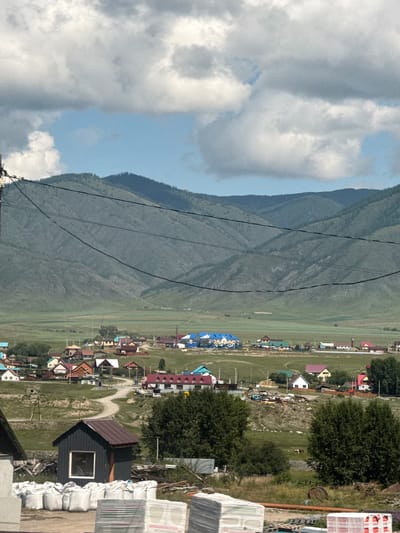 The image depicts a rural landscape near Onguday, Russia, featuring a village nestled against a backdrop of rolling, tree-covered mountains under a cloudy sky. In the foreground, a small, dark shed with a brown roof and a single window is visible. In front of the shed, stacks of white bags, likely containing construction materials, are arranged on the ground. Further into the scene, a winding dirt road leads through the village, which is characterized by a collection of houses with colorful roofs, ranging from blue and red to brown and white. Some houses appear to be under construction. The village is spread across a green, grassy plain that extends towards the mountains. In the mid-ground, two cows graze in a field. Utility lines are strung across the sky in the middle ground. The overall environment suggests a remote, possibly agricultural, settlement.