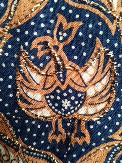 A textile features a brown and blue pattern with a stylized bird motif. The bird is outlined with a thin, glittering gold thread. The background is blue with small white dots, while the bird design alternates between brown and blue with a similar dot pattern. The textile's texture appears woven and slightly textured.