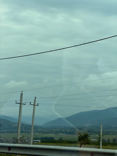 A landscape shot, taken near Tserovani, Georgia, depicts a partly cloudy sky traversed by several power lines attached to concrete utility poles. In the distance, mountains form a backdrop to a valley with green fields and some structures, potentially houses or buildings. A section of a guardrail is visible in the foreground.