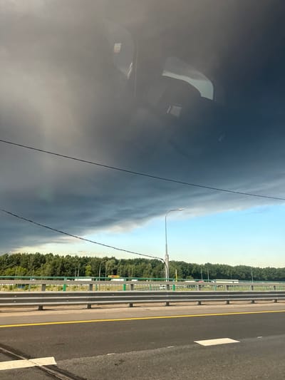 This is a photograph taken from inside a vehicle, looking out at a highway and a dramatic sky. The foreground shows a portion of the asphalt road with a yellow dividing line and a white road marking. Beyond the road is a metal guardrail with a green railing on top, separating the road from a tree-lined area. In the background, a dense line of green trees stretches across the horizon, with streetlights visible amongst them. The sky is predominantly dark and ominous, with heavy, grey storm clouds covering the upper half of the frame. A thin sliver of bright blue sky is visible towards the horizon, contrasting with the approaching storm. Two thin black wires are strung across the sky. The reflection of the vehicle's interior, including a headrest and a window, is visible in the top portion of the image. The scene appears to be captured on a highway, possibly on the outskirts of Yam, Russia, given the presence of the trees and the highway infrastructure. There are no people visible in the image. The overall impression is one of a pending storm.