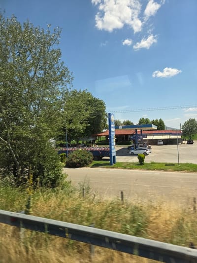 The image depicts a gas station and its surroundings in Klokotos, Greece, on a clear, sunny day. The gas station features a red-tiled roof over the pumps and a blue signpost. Several cars are visible, including a white hatchback at the pump and other vehicles parked. A truck with a load of red cylindrical objects, possibly gas canisters, is parked to the left of the gas station. Trees and green vegetation are scattered throughout the scene, with a large tree in the foreground on the left. A road runs in front of the gas station, and a guardrail is present in the lower portion of the image, suggesting the photo was taken from a moving vehicle. The sky is blue with scattered white clouds.