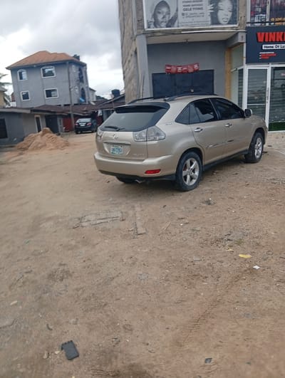 The image depicts a dusty, outdoor scene in Rumuepirikom, Nigeria. A gold-colored Lexus RX 330 SUV is parked on a dirt lot. The vehicle has a license plate that reads "LKM-72960". In the background, a multi-story grey building with an orange roof is visible, as well as a partially constructed building with a corrugated metal roof. Another dark-colored car is parked behind the Lexus. A pile of sand is present to the left of the Lexus. A billboard with images of women is displayed on the side of a building to the right. The ground is covered in dirt, gravel, and debris, with tire tracks visible. The sky is overcast.