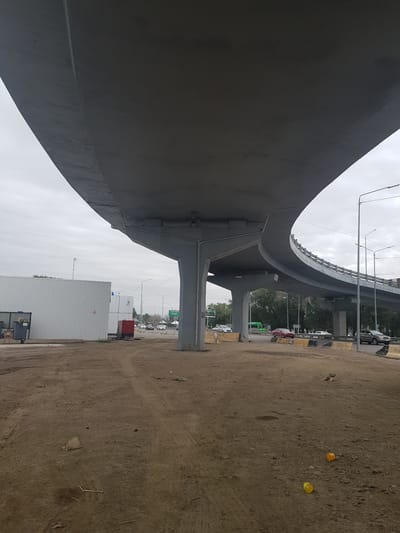 The image shows an underpass of a highway in Бұлақты, Kazakhstan. The underpass is made of concrete and appears to be under construction or recently completed. The ground is unpaved and dirt. There are several vehicles on the highway, including cars and a bus. A white industrial building is visible on the left side of the image. The sky is overcast.