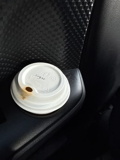 The image shows a disposable coffee cup lid resting inside a vehicle. The lid is white and has a small hole for drinking with a stain around it. Markings on the lid indicate the presence of sugar. The lid rests on black interior trim of the vehicle. Part of the car’s interior has a patterned black texture. The location is unknown; there is no information in the image to definitively determine that the location is Ruma Municipality, Serbia.
