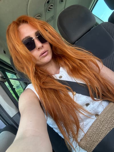 A woman with long, wavy, reddish-orange hair is captured from a slightly high-angle, looking down at the camera, in what appears to be the interior of a car. She is wearing dark sunglasses, a white, eyelet-detailed top, and a straw-like clutch bag is resting on her lap. The background shows a car seat and a glimpse of the outdoors through the car window, suggesting a sunny day. The woman's expression is relaxed, with her lips slightly parted. The scene conveys a casual, on-the-go feel. The location is identified as Rijeka Reževići, Montenegro.