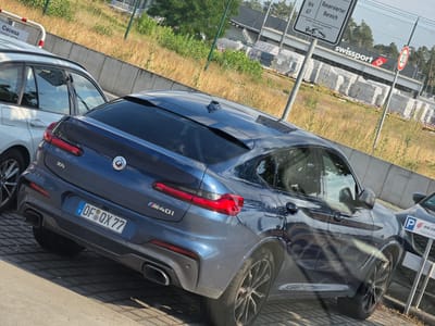 The image shows a dark blue BMW X4 M40i parked outdoors, possibly in a parking lot. The car is seen from the rear, with its taillights and license plate clearly visible. The license plate reads "OF QX 77", with German and European Union insignia. The car has a sporty appearance with dual exhaust pipes and an "M40i" badge.
In the background, a white car is parked next to the BMW. A chain-link fence separates the parking area from a grassy embankment with some vegetation. Behind the fence, there's a sign that says "swissport" with a red logo and "AIR CH". Another sign with "30" inside a red circle suggests a speed limit. Further in the background, there are some buildings and trees.
The lighting suggests it is daytime, with bright sunlight casting shadows. The overall scene appears to be a typical parking area at an airport or a similar transportation hub, given the "swissport" branding, which is an aviation ground handling company. There are no people visible in the image. The weather appears to be clear and sunny. There are no visible emotions as there are no people.