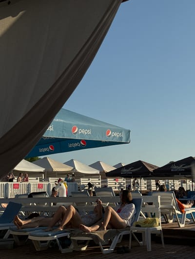 The image depicts a scene at a beach or poolside resort in Luka-Meleshkivska, Ukraine. In the foreground, two people are relaxing on lounge chairs, one with their legs crossed and wearing a white shirt over swimwear, and another with their legs extended. Several lounge chairs are visible, some occupied by other individuals. Overhead, large beach umbrellas with "Pepsi" logos provide shade. In the background, more white and dark umbrellas are set up, some displaying "Belgian Legend" branding. A unicorn inflatable is propped up near a fence in the mid-ground, and several white beach cabanas are lined up behind the lounge chairs. The sky is clear and blue, and the lighting suggests it is late afternoon.
